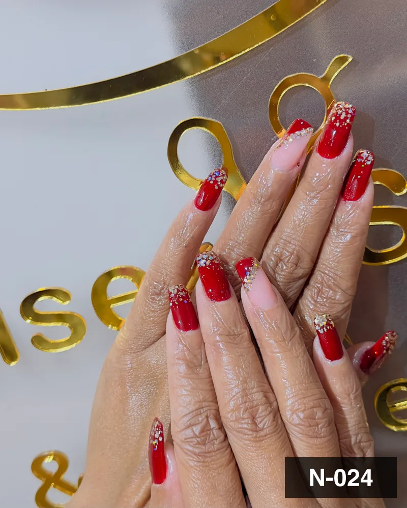 A red long almond set with a gold glitter french edge and crystal detail, against the monogram at Salon By Babita, Haldwani — reference N-024