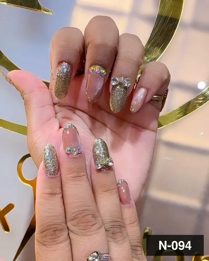 A gold glitter and nude nail set with rhinestone bow charms held against a gold monogram at Salon By Babita, Haldwani — reference N-094