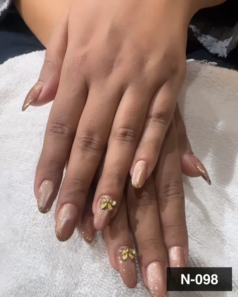 A nude nail set with gold glitter french tips and gold bow charms on both hands at Salon By Babita, Haldwani — reference N-098