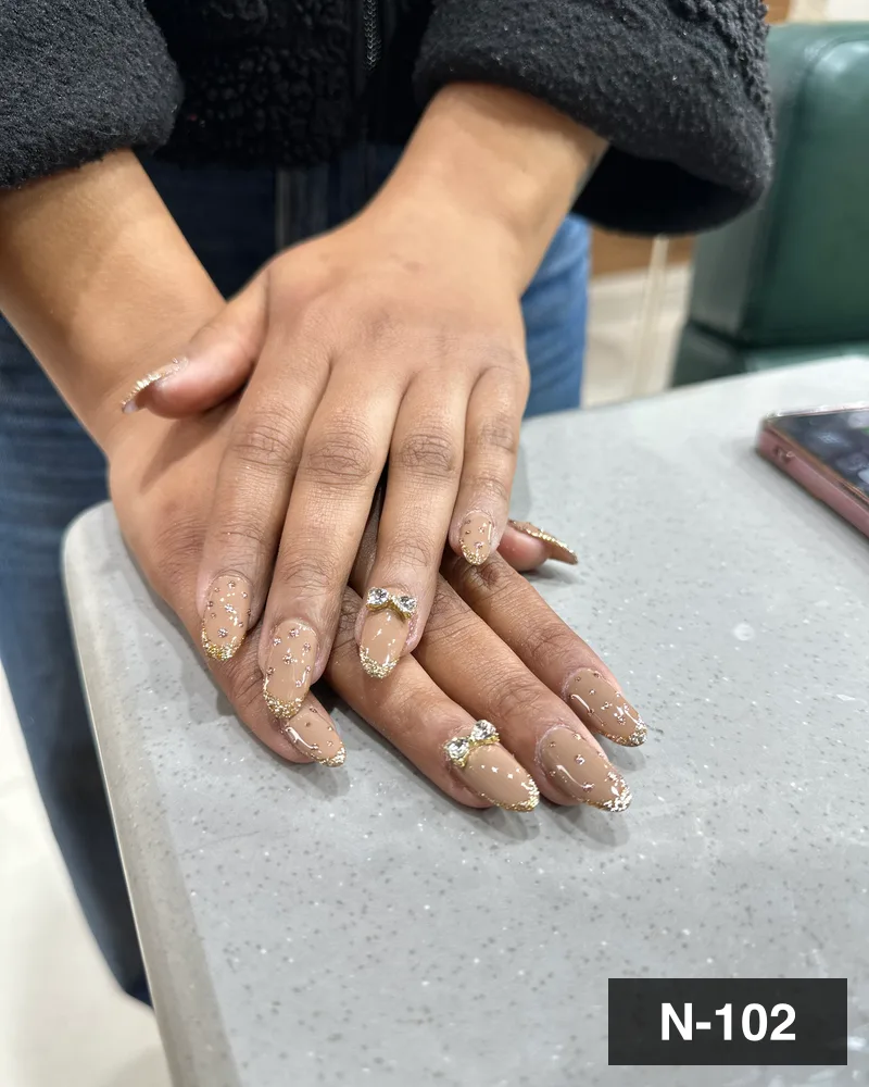 A nude almond nail set with gold glitter french tips and rhinestone bow accents on both hands at Salon By Babita, Haldwani — reference N-102