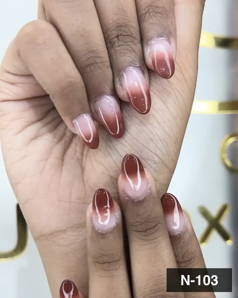 A nude-to-brown ombré almond nail set on both hands against gold lettering at Salon By Babita, Haldwani — reference N-103